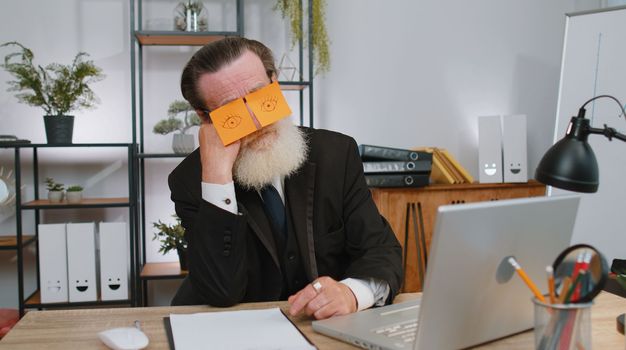 Inefficient tired senior businessman working sleeping on laptop computer with eyes stickers on face at office workplace desk. Elderly lazy manager freelancer man. Business old people cheating to sleep