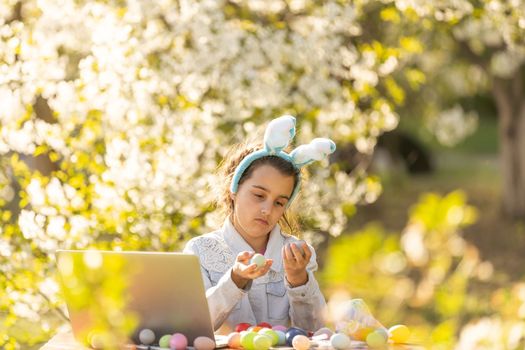 teenage girl paints Easter eggs with a laptop in the garden.