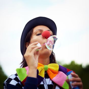 Make the bubble bigger. a clown blowing bubbles at an outdoor festival
