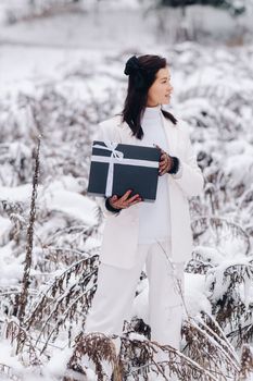 A stylish woman with a white suit with a New Year's gift in her hands in a winter forest. A girl in nature in a snowy forest with a gift box.