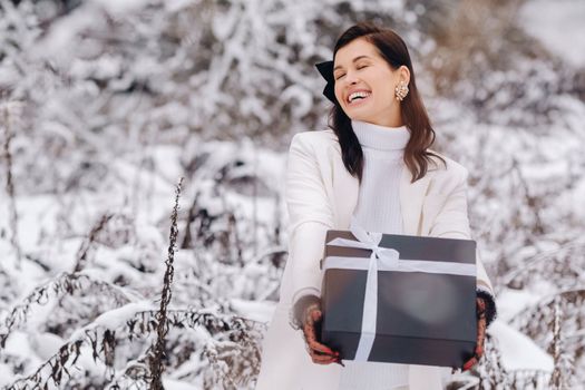 A stylish woman with a white suit with a New Year's gift in her hands in a winter forest. A girl in nature in a snowy forest with a gift box.