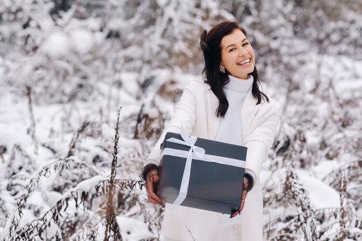 A stylish woman with a white suit with a New Year's gift in her hands in a winter forest. A girl in nature in a snowy forest with a gift box.