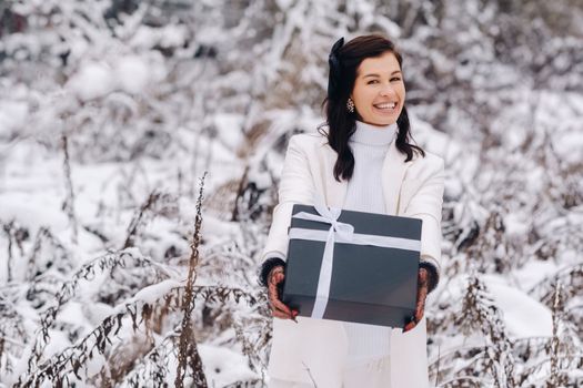 A stylish woman with a white suit with a New Year's gift in her hands in a winter forest. A girl in nature in a snowy forest with a gift box.