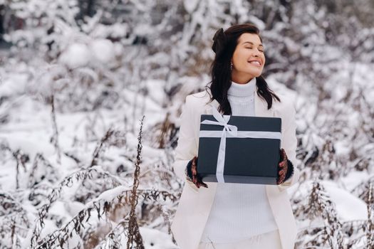 A stylish woman with a white suit with a New Year's gift in her hands in a winter forest. A girl in nature in a snowy forest with a gift box.