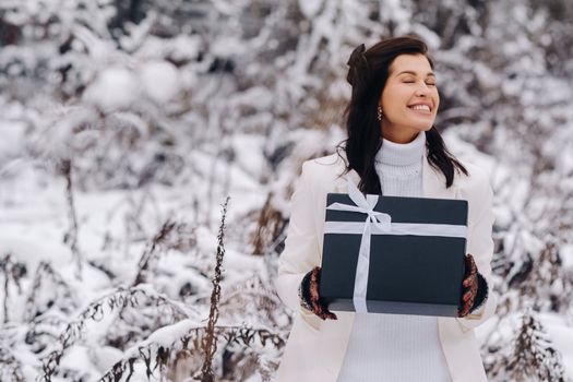 A stylish woman with a white suit with a New Year's gift in her hands in a winter forest. A girl in nature in a snowy forest with a gift box.