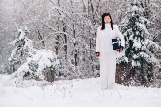 A stylish woman with a white suit with a New Year's gift in her hands in a winter forest. A girl in nature in a snowy forest with a gift box.