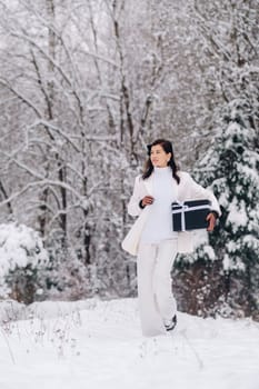 A stylish woman with a white suit with a New Year's gift in her hands in a winter forest. A girl in nature in a snowy forest with a gift box.