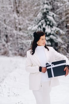 A stylish woman with a white suit with a New Year's gift in her hands in a winter forest. A girl in nature in a snowy forest with a gift box.