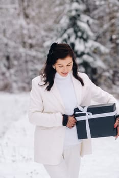 A stylish woman with a white suit with a New Year's gift in her hands in a winter forest. A girl in nature in a snowy forest with a gift box.