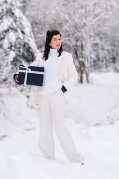 A stylish woman with a white suit with a New Year's gift in her hands in a winter forest. A girl in nature in a snowy forest with a gift box.