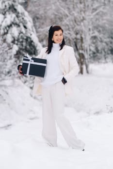 A stylish woman with a white suit with a New Year's gift in her hands in a winter forest. A girl in nature in a snowy forest with a gift box.