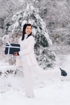 A stylish woman with a white suit with a New Year's gift in her hands in a winter forest. A girl in nature in a snowy forest with a gift box.