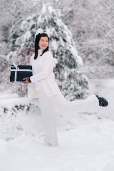 A stylish woman with a white suit with a New Year's gift in her hands in a winter forest. A girl in nature in a snowy forest with a gift box.