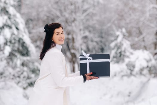 A stylish woman with a white suit with a New Year's gift in her hands in a winter forest. A girl in nature in a snowy forest with a gift box.