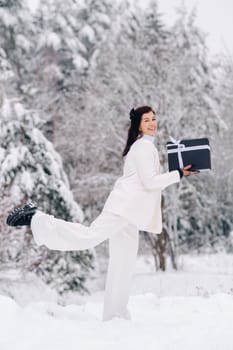 A stylish woman with a white suit with a New Year's gift in her hands in a winter forest. A girl in nature in a snowy forest with a gift box.