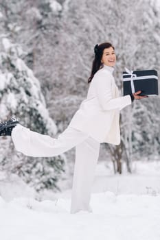 A stylish woman with a white suit with a New Year's gift in her hands in a winter forest. A girl in nature in a snowy forest with a gift box.