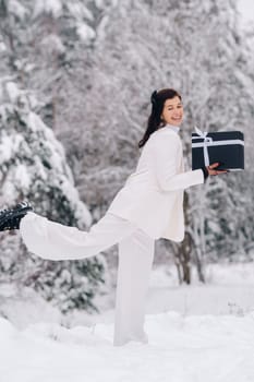 A stylish woman with a white suit with a New Year's gift in her hands in a winter forest. A girl in nature in a snowy forest with a gift box.