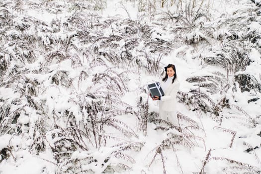 A stylish woman with a white suit with a New Year's gift in her hands in a winter forest. A girl in nature in a snowy forest with a gift box.