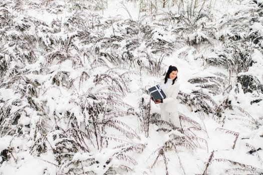 A stylish woman with a white suit with a New Year's gift in her hands in a winter forest. A girl in nature in a snowy forest with a gift box.