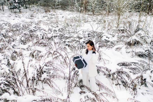 A stylish woman with a white suit with a New Year's gift in her hands in a winter forest. A girl in nature in a snowy forest with a gift box.