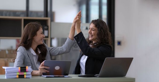 Two young Asian businesswomen show joyful expression of success at work smiling happily with a laptop computer in a modern office..