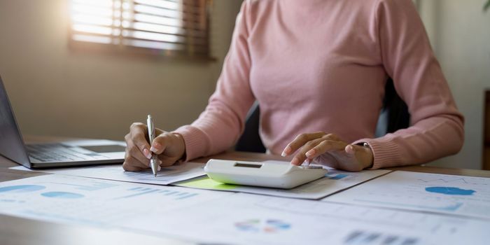 Close up of businesswoman or accountant hand holding pen working on calculator to calculate business data, accountancy document and laptop computer at office, business concept.