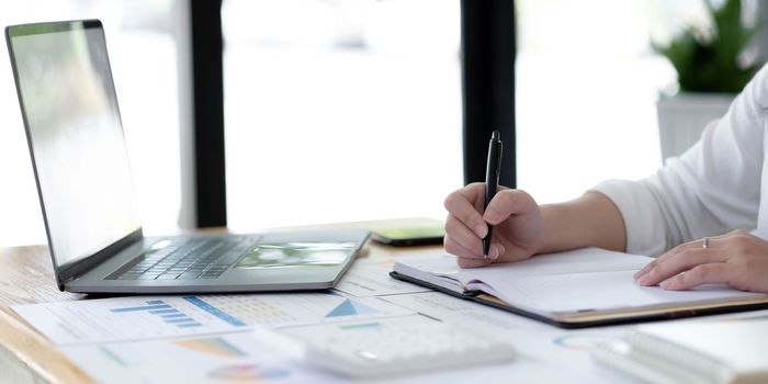 Young business woman sitting at table and taking notes in notebook.On table is laptop, smartphone.