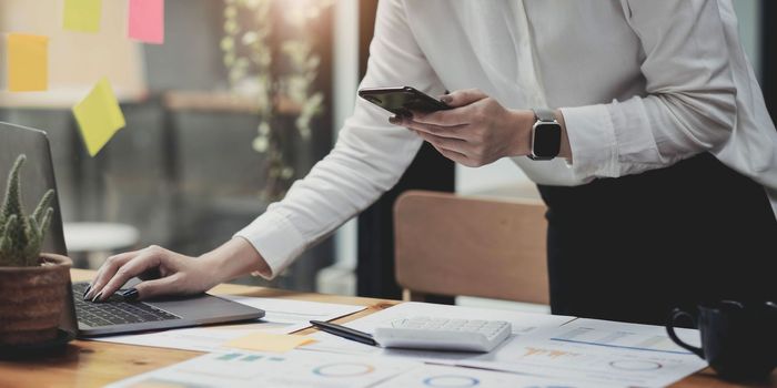 Young accountant hand holding smartphone to call marketing consultants and using laptop computer to analyze sales growth in the global workplace market. accounting concept.