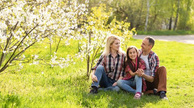 Outdoor portrait of happy young family playing in spring park under blooming tree, lovely couple with little child having fun in sunny garden.