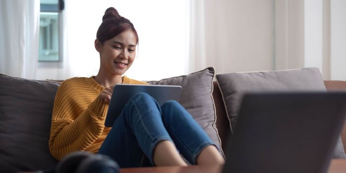 Candid of asian woman using tablet for watching online movie stream mobile device on sofa at living room. Young happy girl enjoying with entertainment media online on weekend..