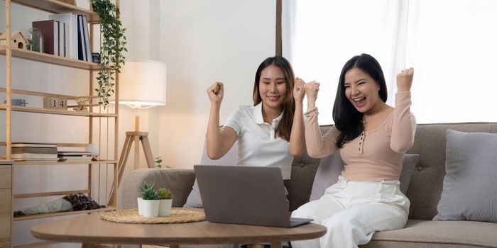 Exciting young asian women LGBT lesbian happy couple sitting on sofa using laptop a computer and phone in living room at home. LGBT lesbian couple together.