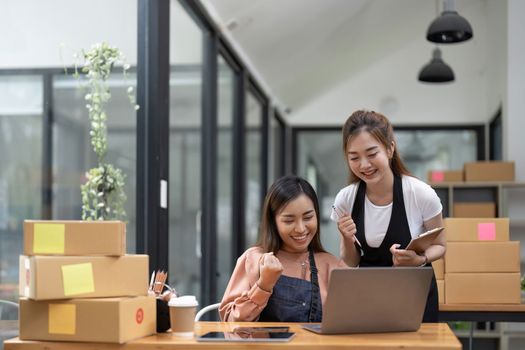 Happy two asian women entrepreneur, Smile for sales success after checking order from online shopping store in a smartphone at home office, Concept of merchant business online and eCommerce.
