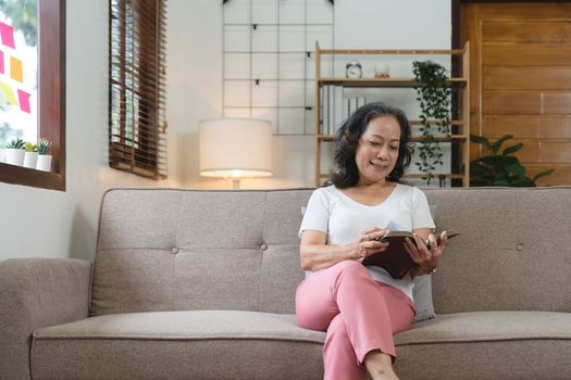smiling attractive retired woman reading book while sitting on sofa at home