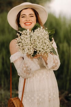 Portrait of a beautiful woman in a white dress and a hat with lilies of the valley at sunset. A girl in nature. Spring flowers.