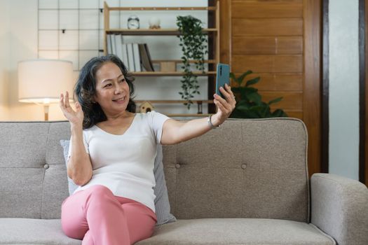 Asian elderly woman taking selfie on mobile phone while sitting on sofa and smiling.