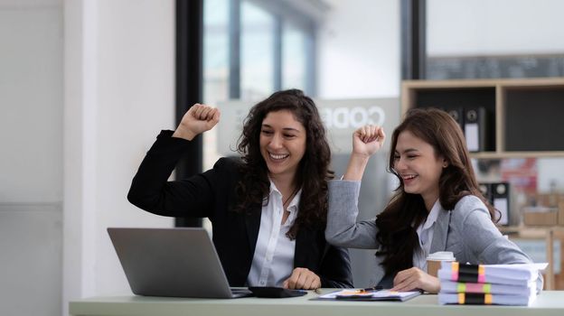 Two young Asian businesswomen show joyful expression of success at work smiling happily with a laptop computer in a modern office..