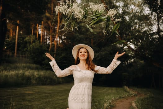 Portrait of a beautiful woman in a white dress and hat tossing lilies of the valley. A girl in nature. Spring flowers.