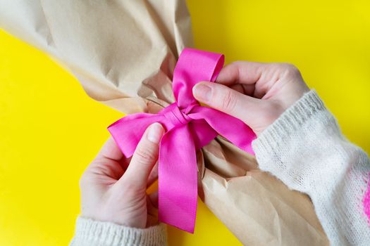 Top view of female hands tying a bouquet with a pink ribbon with gifts on a yellow background. Close-up, space for an inscription