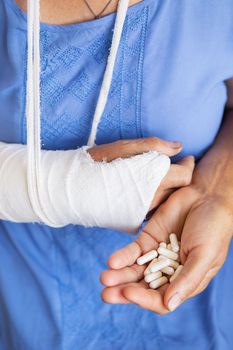 A senior retired woman with a rewound arm in a cast and a bandage holds anesthetic pills. Blow, fracture, bones, hospital