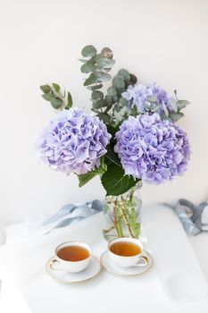 Two cups of black tea in a porcelain beautiful white cup with a gold cut. Beautiful bouquet of purple hydrangea on the table. Tea break