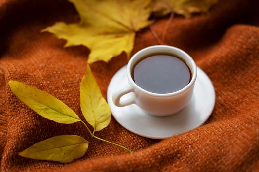 Still life with black coffee in a white cup that stands on a warm cozy sweater, top view, decorations with yellow leaves on the background