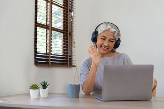 Smiling senior asian woman with headphones on her head sitting at a table in front of a laptop and greeting family during quarantine