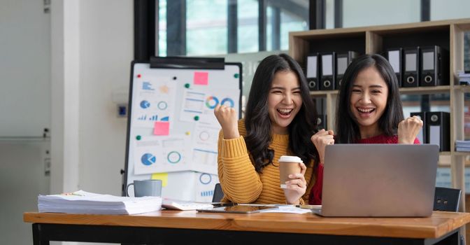 Two young Asian businesswomen show joyful expression of success at work smiling happily with a laptop computer in a modern office..