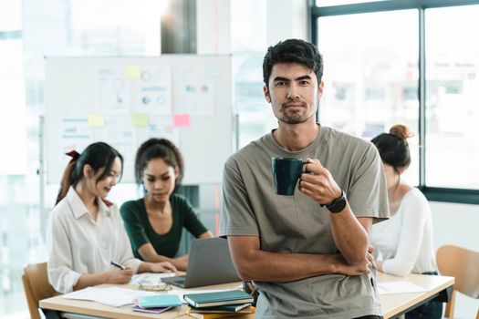Portrait of happy asian small business owner posing with hands folded. Millennial male team leader smiling, looking at camera, employees working in modern office behind.