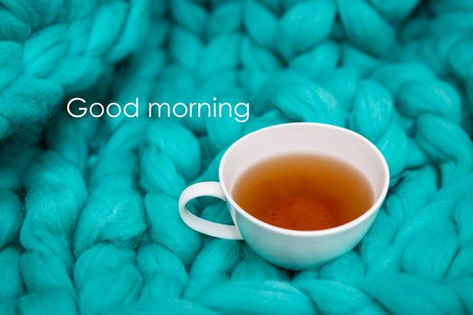 The concept of coziness and comfort is a green knitted blanket on which there is a white cup of tea. Close-up. Good morning lettering