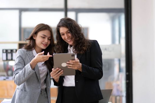 Two beautiful Asian businesswomen standing using digital tablet consulting and analyzing information in office work..