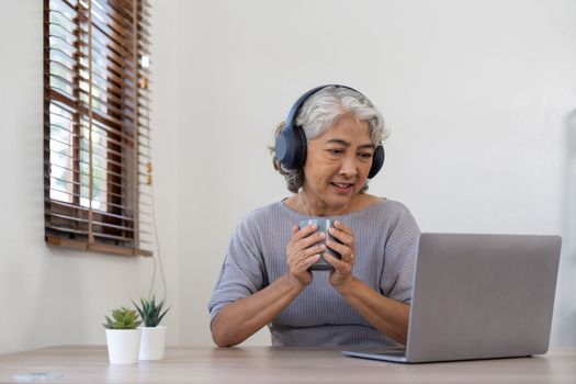 Senior woman using laptop while wearing headphones at home - Joyful elderly lifestyle and technology concept.