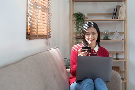 Beautiful young woman holding credit card while using laptop computer and mobile phone sitting at the living room. Black friday online shopping concept.