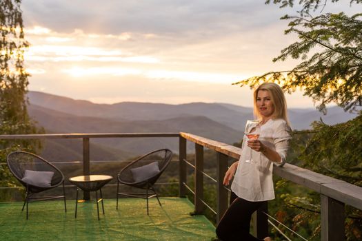 Woman holding glass of white wine across beautiful mountains landscape and sunset.