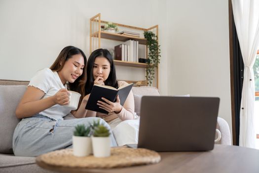 Two young asian girls are sitting on couch and reading book at home.
